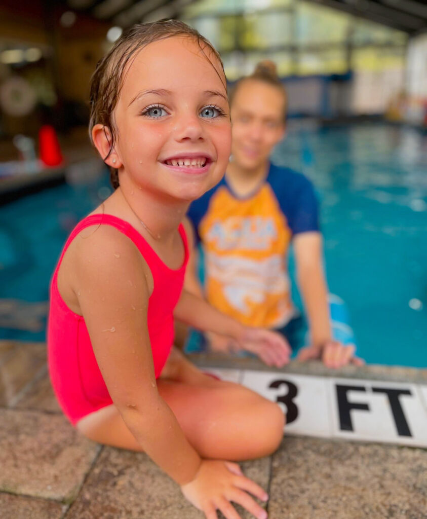 A little girl sitting next to an adult in the pool.